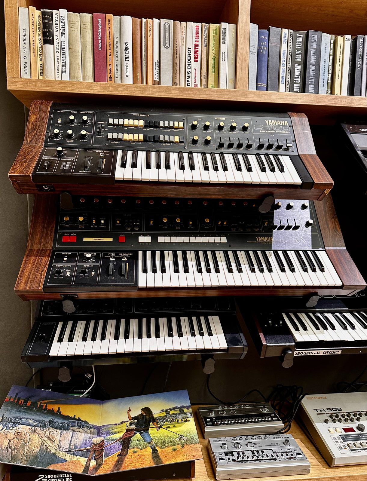 Stack of vintage synthesizers on wooden shelves under a library.
